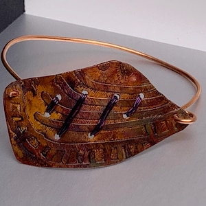 Puede incluir: Una pulsera de color cobre con una pieza central en forma de hoja. La pieza central tiene una superficie texturizada y estampada con costuras moradas oscuras y detalles blancos. La pulsera es un simple alambre de cobre.