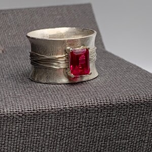 May include: A silver ring with a wide band and a rectangular, bright red gemstone. The band has a textured finish and is accented with thin, wrapped wire. The gemstone is set in the center of the ring.