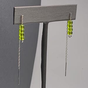 May include: A pair of dangle earrings featuring a chain and a cluster of four round, lime green beads. The earrings are suspended from a silver-colored chain and have a long, thin, silver-colored bar at the end.