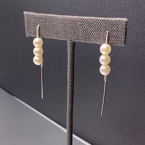 May include: A pair of elegant earrings featuring three creamy white pearls suspended from a delicate silver chain. A long, slender silver bar hangs below the pearls, creating a modern and sophisticated look. The earrings are displayed on a gray fabric earring stand.