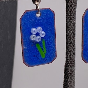 May include: A pair of blue and green enamel earrings with a white flower design. The earrings are rectangular in shape and have a silver hook.