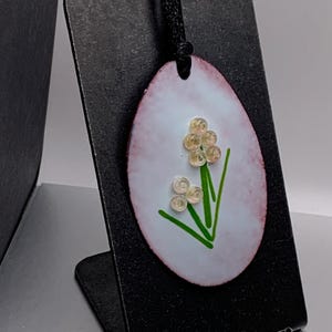 Puede incluir: Un colgante ovalado blanco con una flor verde y cuentas blancas. El colgante está en un cordón negro y está colgado de un soporte de exhibición negro.