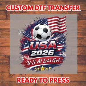 May include: A DTF transfer design featuring a soccer ball, the American flag, and the text "USA 2026" with "U-S-A! Let's Go!" and "READY TO PRESS". The design incorporates red, white, blue, and gold colors.