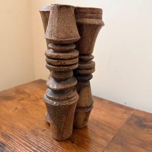 May include: A brown ceramic sculpture featuring two conjoined vertical columns with a textured, speckled surface. Each column displays a series of stacked, rounded, and flared geometric segments, creating a rhythmic, layered appearance in earthy tones.