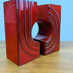 Könnte beinhalten: Zwei rote Keramik-Dekorationsobjekte mit geometrischen Designs. Eines hat vertikale Linien, das andere hat gebogene Linien. Beide haben eine glänzende Oberfläche und sind vor einem hellblauen Hintergrund platziert.