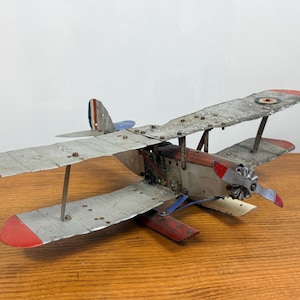 Op de afbeelding: Een metalen dubbeldekker model in vintage stijl met een verweerde zilveren afwerking en rood gekleurde vleugeltips. Het vliegtuig is voorzien van een propeller aan de voorzijde, een rood rompdeel en een staartvin met blauw-wit-rode strepen.