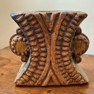 May include: A brown ceramic vase with a textured surface featuring an hourglass shape. The exterior is decorated with vertical rows of raised, bead-like patterns and circular accents on the sides, finished in a matte, earthy brown glaze.