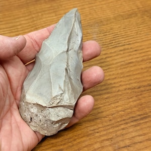 May include: A gray, hand-held stone tool with a rough, textured surface. The tool has a pointed end and a wider, rounded base. The stone appears to be chipped and shaped, possibly for cutting or scraping. The background is a wooden surface.