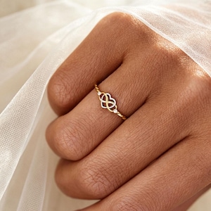 May include: A thin gold ring featuring a central heart-shaped motif intertwined with an infinity symbol. The design includes two small, clear, round-cut stones set on either side of the central heart element, mounted on a delicate, polished gold band.