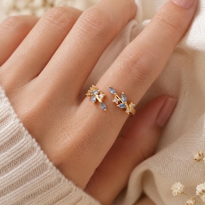May include: A gold-toned open ring featuring two branch-like ends adorned with small, marquise-cut light blue stones and tiny clear round accent stones. The design mimics a delicate botanical motif, with the stones set along the metal branches.