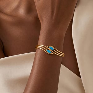 May include: A gold-toned cuff bracelet featuring three parallel bands that curve to form an open space. Two marquise-shaped, bright blue stones are set within the center of the design, positioned horizontally along the top of the wrist.