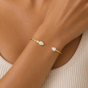 Puede incluir: Una pulsera abierta de tono dorado con dos piedras de ópalo iridiscentes en forma de lágrima en cada extremo. La pulsera tiene una banda de metal lisa y pulida que se curva alrededor de la muñeca, mostrando un diseño sencillo con gemas.