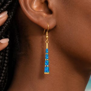 Puede incluir: Un pendiente colgante de tono dorado con un diseño de barra vertical delgada y cónica. El pendiente está incrustado con cuatro segmentos distintos de material azul vibrante similar al ópalo, separados por finas bandas de metal dorado.
