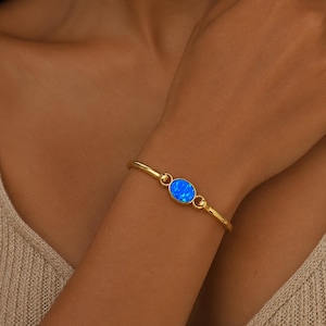 May include: A gold-toned bangle bracelet featuring a central oval-shaped blue opal stone. The bracelet has a smooth, polished metal band that connects to the bezel-set stone, creating a continuous circular design around the wrist.