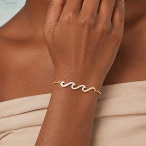 May include: A gold-toned chain bracelet featuring a central wave-shaped charm. The wave charm is filled with a white, iridescent material that reflects light, and the bracelet is secured with a thin, delicate gold-coloured metal chain.