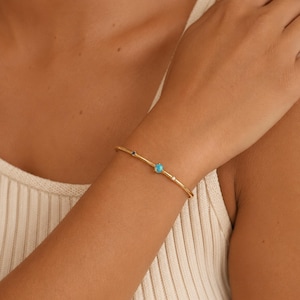 May include: A thin gold bangle bracelet featuring three small, round, bezel-set gemstones: a dark blue stone, a bright turquoise stone in the center, and a small white stone, all spaced evenly along the polished gold band.