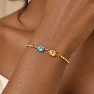 Könnte beinhalten: Ein goldenes Armband mit zwei oval geschliffenen Edelsteinen, einem blauen und einem gelben, die in eine feine Metallkette eingefasst sind. Das Armband zeichnet sich durch eine glatte, polierte Oberfläche und einen einfachen Verschluss aus.