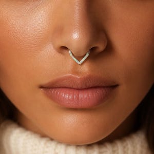 May include: A V-shaped septum ring featuring a metallic silver-toned frame with an iridescent, white-toned inlay. The jewelry is positioned in the center of the nose, showcasing a clean, angular design that follows the contour of the nasal septum.