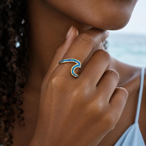 May include: A gold ring shaped like a stylized ocean wave, featuring a vibrant blue inlay that follows the curve of the wave design. The ring is worn on a finger, showcasing the metallic gold band and the bright blue textured surface.
