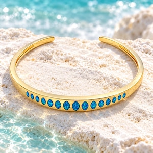 May include: A gold-toned open cuff bracelet featuring a row of eleven marquise-cut blue opal-style stones set along the center. The metal band has a smooth, polished finish and a curved design that tapers slightly toward the open ends.