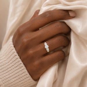 Puede incluir: Un anillo de oro con tres piedras de ópalo blanco iridiscente redondas dispuestas en fila. La piedra central es ligeramente más grande que las dos piedras laterales, todo montado sobre una fina banda de oro pulido en el dedo.