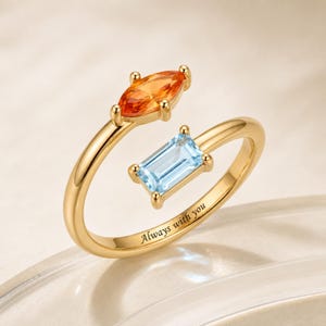 Pode incluir: Um anel aberto de ouro com uma pedra preciosa laranja de corte marquise e uma pedra azul-clara de corte retangular nas extremidades. A banda está gravada com o texto Always with you numa fonte serifada preta e acabamento polido.