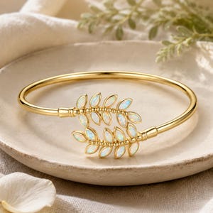 Puede incluir: Una pulsera rígida de tono dorado con dos ramas simétricas en forma de hoja que se encuentran en el centro. Cada hoja está incrustada con una piedra de ópalo iridiscente de corte marquesa, creando un diseño botánico con acabado pulido.