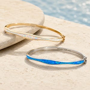 May include: Two hinged bangle bracelets, one gold-toned with a white iridescent inlay and one silver-toned with a blue iridescent inlay. Both bracelets feature the engraved text Always with you on the inner band and a secure clasp mechanism.