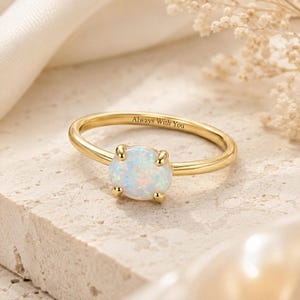 May include: A gold ring featuring a central oval-shaped iridescent opal stone held by four prongs. The inner band of the ring is engraved with the text Always With You in a clear, dark font, contrasting against the polished gold metal surface.