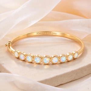 May include: A gold-toned hinged bangle bracelet featuring a row of nine round, iridescent white opal-like stones set in individual bezels. The inner band is engraved with the text Always with you, and the piece includes a secure side clasp mechanism.