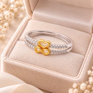 Puede incluir: Un anillo de tono plateado con un diseño de doble banda y una hilera de pequeñas piedras transparentes a cada lado. El centro del anillo muestra una forma de corazón en tono dorado apilada sobre un símbolo de infinito en tono dorado.