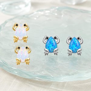 Pode incluir: Dois pares de brincos em forma de sapo. Um par é de metal dourado com pedras brancas iridescentes em forma de gota. O segundo par é de metal prateado com pedras azuis brilhantes e iridescentes em forma de gota.