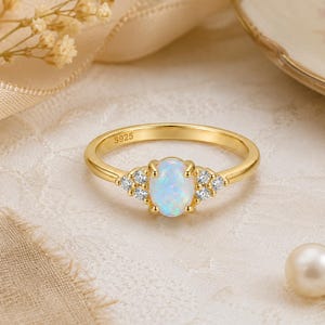 May include: A gold-toned ring featuring a central oval-shaped iridescent opal gemstone held by four prongs. The band is stamped with S925 and includes three small clear round stones set on each side of the central opal in a cluster arrangement.
