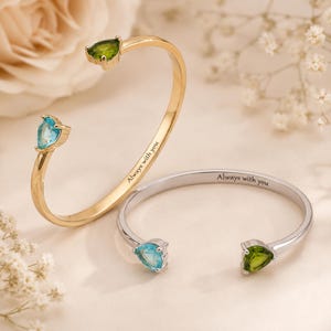 May include: Two open cuff bracelets, one gold-toned and one silver-toned, each featuring a pear-shaped green stone and a pear-shaped blue stone at the ends. The inner band of each bracelet is engraved with the text Always with you in black script.