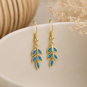 May include: A pair of gold-toned drop earrings featuring a leaf-shaped design. Each earring consists of a thin gold wire stem with five small, oval-shaped blue iridescent stones arranged to resemble a leaf, suspended from a gold hook wire.