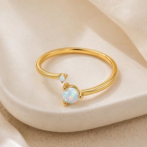 May include: A gold-toned open ring featuring two round, iridescent opal-like stones of different sizes. The larger stone is set at one end of the band, while a smaller stone is positioned at the opposite end, creating a bypass style design.