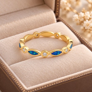 Puede incluir: Un anillo de banda dorada con un patrón alterno de piedras de ópalo azul en talla marquesa y piedras blancas transparentes en talla redonda. El metal tiene un acabado pulido y las piedras están engastadas en biseles individuales alrededor.