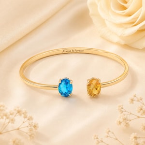 May include: A gold open cuff bracelet featuring an oval-cut blue gemstone on one end and an oval-cut yellow gemstone on the other. The inner band is engraved with the text Always & Forever in a clean, dark font, creating a balanced design.