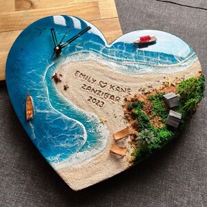 Op de afbeelding: Hartvormige wandklok met een strandscène met blauwe harsgolven, een zandstrand en kleine bootjes. De wijzerplaat bevat de tekst "EMILY & KANE ZANZIBAR 2023". De wijzers zijn zwart.