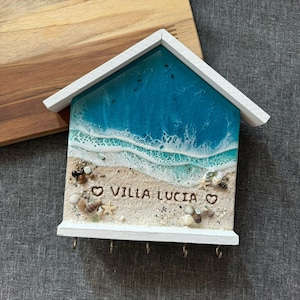 May include: A house-shaped key holder depicting a beach scene. The top section shows a blue ocean design, and the bottom has sand, shells, and the text "VILLA LUCIA". It has a white roof and a row of hooks for keys.