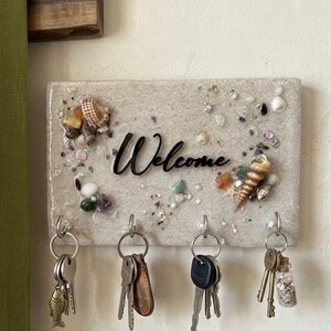 Könnte beinhalten: Ein rechteckiger Schlüsselhalter zur Wandmontage mit einer sandigen, strukturierten Oberfläche, die mit verschiedenen Muscheln, kleinen Steinen und einem zentralen schwarzen Schriftzug Welcome verziert ist. Vier silberne Haken.