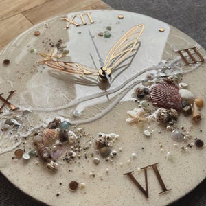Op de afbeelding: Een ronde wandklok met een strandthema. De wijzerplaat heeft een zanderige textuur met ingebedde schelpen, zeesterren en kleine steentjes. Gouden Romeinse cijfers markeren de uren. De wijzers zijn goudkleurig en het ontwerp roept een kustesthetiek op.
