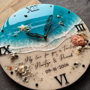 May include: A circular wall clock with a beach theme. The clock face depicts a blue and white ocean scene, adorned with seashells, starfish, and Roman numerals. The black clock hands point to the time, and the text reads "My love for you is Timeless Maritza & Pranah 09-16-2006".