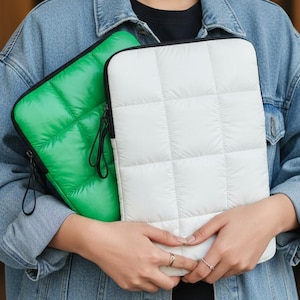 May include: Two padded laptop sleeves, one white and one bright green, are held in front of a person. The sleeves have a quilted pattern and black zippers. The person is wearing a denim jacket.