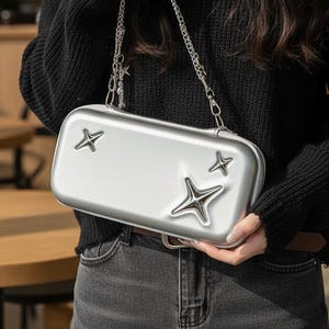 May include: Silver rectangular handbag with star detailing and a chain strap. The bag has a metallic finish and is held by a person wearing a black jumper and jeans. The stars are silver.