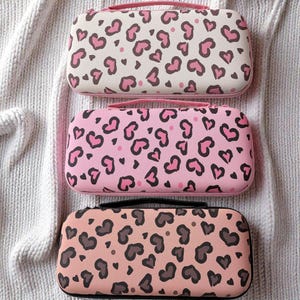 Puede incluir: Tres estuches rectangulares con estampados de leopardo en forma de corazón. Los estuches son en tonos rosa, crema y melocotón, con detalles en negro y rosa. Cada estuche tiene un asa y cremallera a juego.