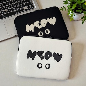 Puede incluir: Dos fundas para portátiles, una negra y otra blanca, apiladas. Cada funda presenta la palabra "MEOW" en letras blancas en negrita, con dos formas de ojos circulares debajo. Las fundas están diseñadas para proteger portátiles.