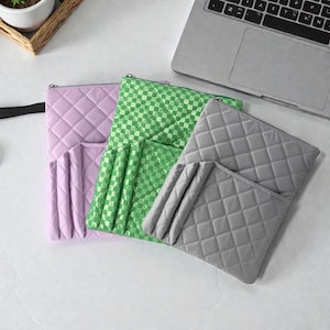 Op de afbeelding: Drie gewatteerde ritszakjes in pastelkleuren. De zakjes zijn in lavendel, groen en grijs. Het lavendelkleurige zakje heeft een ruitpatroon, het groene zakje heeft een ruitpatroon en het grijze zakje heeft een ruitpatroon. Elk zakje heeft een zak aan de voorkant.