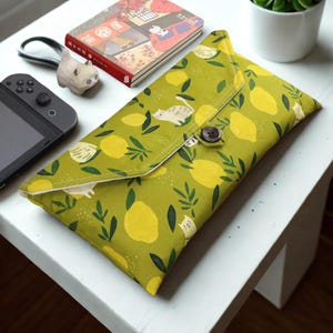 May include: A green fabric case with a lemon and cat pattern, secured with a button closure. The case is rectangular and appears to be for a handheld gaming device. Other items on the white surface include a small cat figurine, a notebook, and a potted plant.