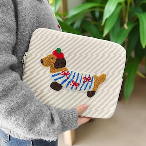 May include: Cream-colored, corduroy laptop sleeve with a zipper closure. The sleeve features an embroidered dachshund wearing a red hat, blue and white striped shirt, and red bows. The dog has brown, black, and tan details.